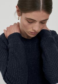 Navy gestrickter Pullover mit texturiertem Finish, rundem Ausschnitt und gerippten Bündchen. Das Modell hat die Hände auf den Schultern und zeigt eine Nahaufnahme.