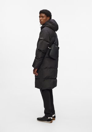 Black puffer coat with a hood, featuring a zip closure and a sleek design. Worn with black pants and sporty shoes; a small black bag is attached.