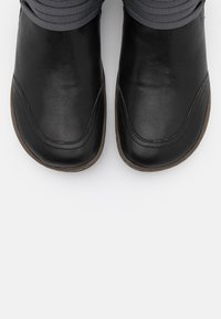 Czarne skórzane buty z zaokrąglonymi czubkami, przeszyciami dekoracyjnymi oraz elastycznymi szarymi wstawkami, wyposażone w płaską gumową podeszwę.