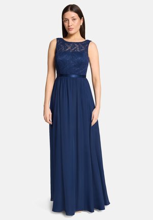 Žena oblečená v tmavomodrých večerných šatách bez rukávov s čipkovaným živôtikom a saténovým opaskom, stojaca na svetlom, jednoduchom pozadí.