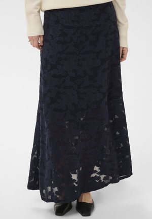 Kobieta w długiej granatowej koronkowej spódnicy w kwiaty, w połączeniu z czarnymi spiczastymi butami i kremowym swetrem, pokazana od talii w dół.