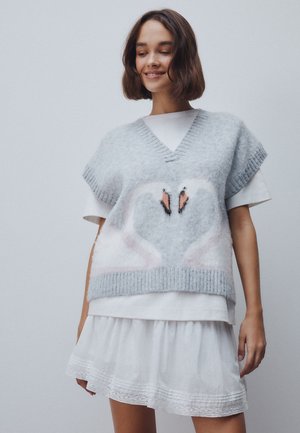 Giovane donna che indossa un maglione smanicato grigio chiaro con due cigni che formano un cuore, sopra una t-shirt bianca e una gonna.
