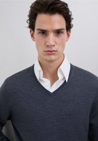 Szary sweter z dekoltem w serek o fakturowanym wykończeniu, noszony na białej koszuli z kołnierzykiem. Z subtelnym logo na lewej piersi.