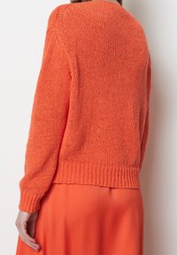 Pomarańczowy sweter z dzianiny o teksturowanym wykończeniu, z okrągłym dekoltem i ściągaczem na dole. Nosi się go na pasującej pomarańczowej spódnicy.