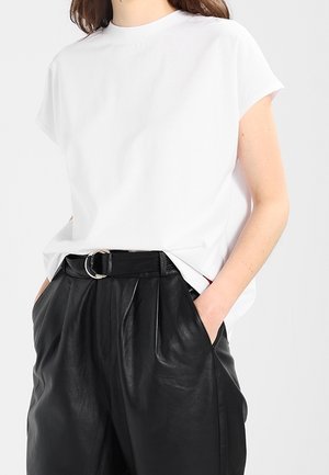 Femme portant un chemisier blanc uni à manches courtes rentré dans un pantalon en cuir noir taille haute avec une ceinture noire. Mains dans les poches.