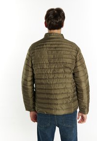 Giacca puffer verde oliva con cuciture orizzontali, colletto alto e texture liscia e lucida. Indossata sopra jeans di denim.
