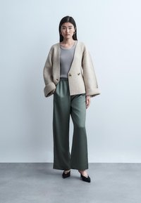 Woman standing against blank wall wearing beige oversized jacket, gray ribbed top, loose green pants, and black heels.