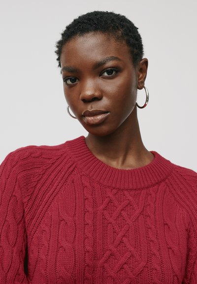 Raudonas megztinis su nertomis akytėmis ir rišamu apykakle. Pasižymi masyviais raštais ir sudėtingomis austomis tekstūromis. Papuoštas auskarais su žiedais.