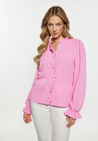 Blusa rosa con un design testurizzato, dotata di colletto arricciato e chiusura con bottoni davanti, abbinata a pantaloni bianchi. Maniche lunghe con polsini arricciati.