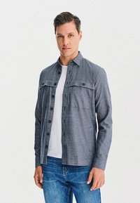 Mężczyzna w niebieskiej koszuli zapinanej na guziki, noszący białą koszulkę i niebieskie dżinsy, stojący na tle jednolitej białej ściany.