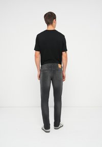 Camiseta negra, jeans ajustados gris con un parche de cuero en la parte posterior de la cintura, y zapatillas negras y blancas, vista desde atrás.
