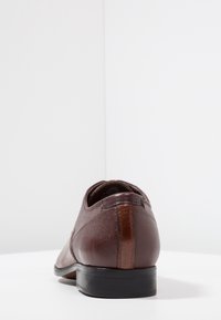 Chaussure en cuir marron avec une finition texturée et des accents lisses ; elle présente une forme arrondie, une semelle noire fine et un détail de talon contrastant.