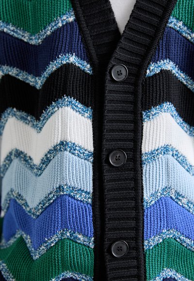 Knitted cardigan featuring a chevron pattern in black, green, blue, white, and light blue, with a ribbed black collar and four buttons.