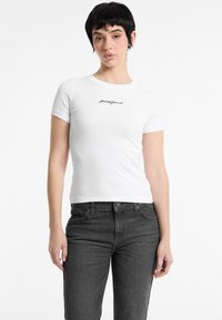 T-shirt blanc ajusté à manches courtes, texture côtelée, et logo "guessjeans" en noir sur le devant, associé à un jean gris foncé.