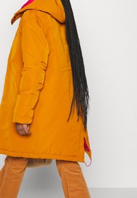 Personne aux longues tresses portant un manteau à capuche orange et un pantalon en velours côtelé marron clair, marchant devant un fond blanc uni.