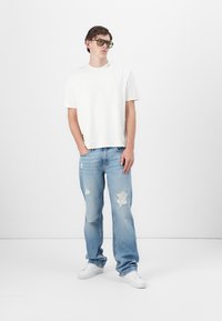 White cotton t-shirt with crew neck, paired with light blue distressed jeans. Model wears green-tinted sunglasses and white sneakers.