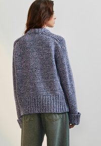 Pletený svetr v purpurových a šedých odstínech, se silnou strukturou, žebrovaným lemem a manžetami a volným střihem. Nosený s zelenými kalhotami.