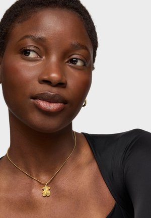 Goldene Halskette mit floralen Anhänger, getragen über einem schwarzen Oberteil. Glatte Hautstruktur und kurze, lockige Haare sind sichtbar.