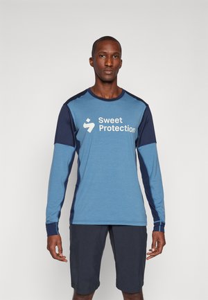 Svetlo modrá košeľa s dlhými rukávmi s tmavomodrými akcentmi a logom "Sweet Protection". Nosí sa s tmavými šortkami, ktoré zdôrazňujú športový dizajn.
