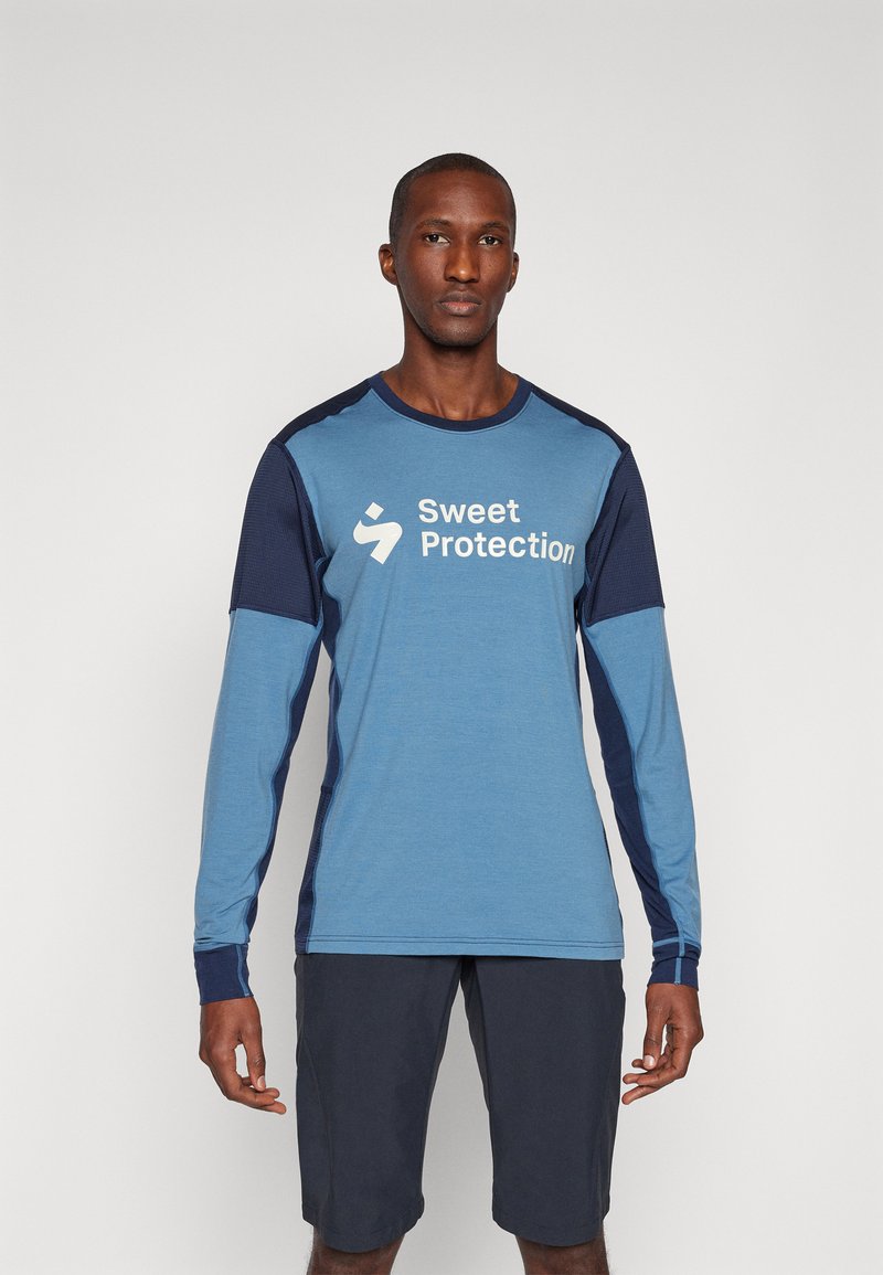 Svijetloplava majica dugih rukava s tamnoplavim detaljima i logom "Sweet Protection". Nosena s tamnim hlačama, prikazujući sportski dizajn.
