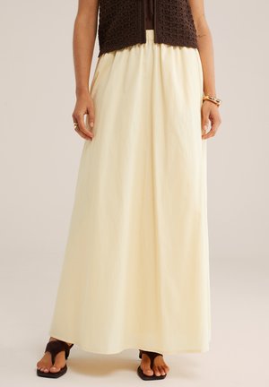 Inimene, kes kannab kreemikaid laiade jalgadega pükse, tumedat heegeldatud vesti, musti sandaale, kuldset käevõru ja sõrmust beeži taustal.