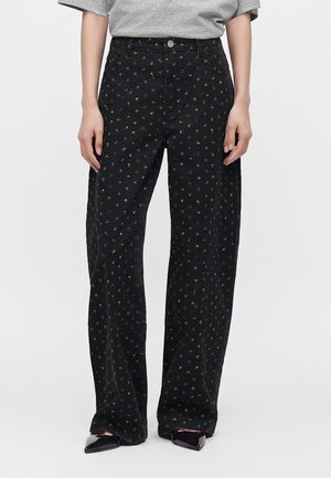 Personne portant un pantalon noir à jambes larges avec de petits motifs floraux colorés et des chaussures noires pointues, associé à un haut gris.