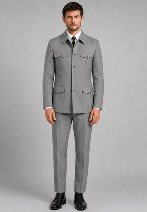 Homme aux cheveux courts et bouclés portant un costume gris sur mesure avec poches boutonnées, chemise blanche, cravate noire et chaussures noires, debout devant un fond gris.