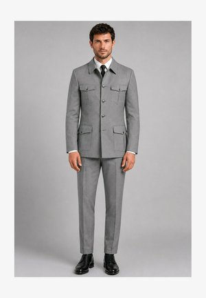 Homme aux cheveux courts et bouclés portant un costume gris sur mesure avec poches boutonnées, chemise blanche, cravate noire et chaussures noires, debout devant un fond gris.