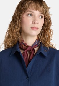 Camicia blu navy con tessuto testurizzato e un piccolo logo. Abbinata a una sciarpa bordeaux con un motivo, annodata attorno al collo.