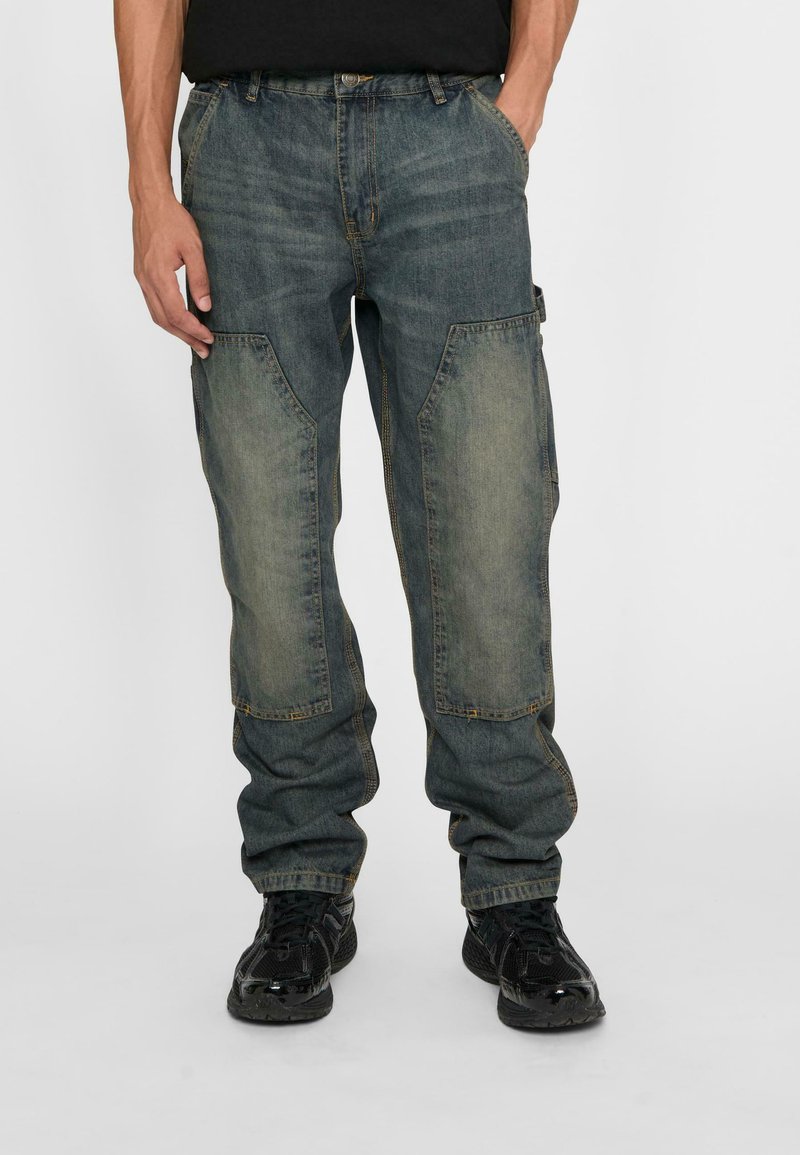 Blue denim cargo pants with light-colored knee patches, loose fit, and pockets on the sides, paired with black sneakers.