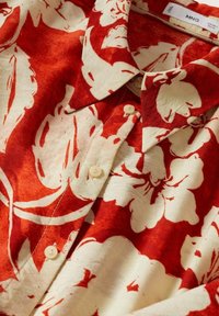 Camisetta a maniche corte rossa e crema con motivo floreale, dotata di colletto a punta, chiusura con bottoni e tessuto leggero.