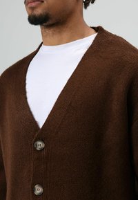 Pull marron avec une finition douce et texturée. Il présente un décolleté en V profond, trois boutons et est superposé à un t-shirt blanc.