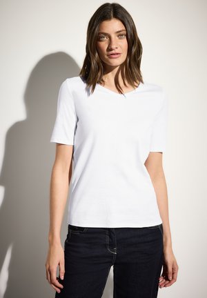 Witte kortekorte mouw V-hals t-shirt, gemaakt van een lichte katoenmix, gecombineerd met donkerblauwe spijkerbroek, staande tegen een lichte achtergrond.