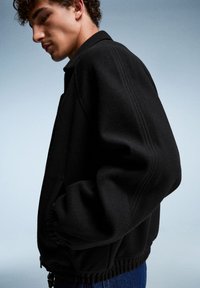 Veste en polaire noire avec une coupe décontractée, des poignets côtelés et un ourlet élastique. Présente un col classique et des poches latérales. Le fond est bleu clair.