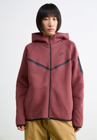 Pessoa com cabelo preto curto, vestindo uma sweatshirt Nike marrom com zíper e detalhes pretos, e calças bege, de pé contra um fundo branco.