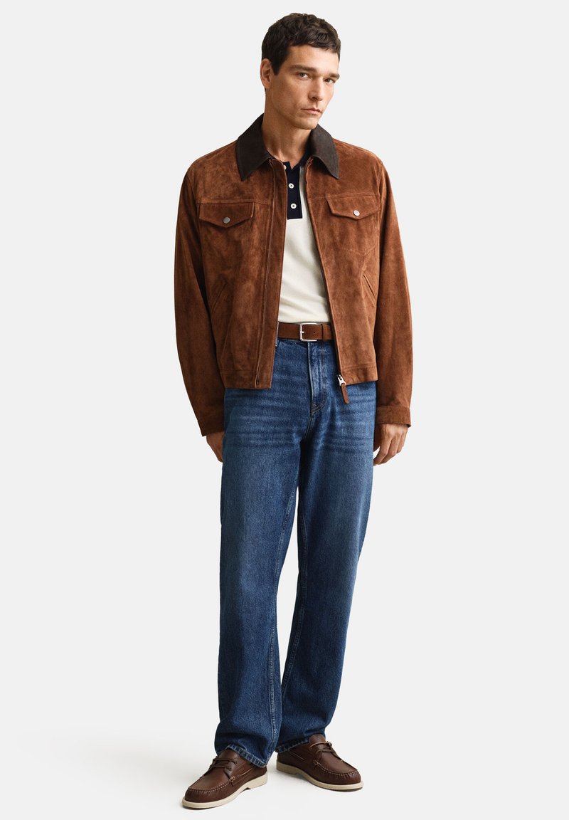 Giovane uomo con giacca in camoscio marrone, polo bianca e nera, jeans blu e scarpe in pelle marrone, in piedi davanti a uno sfondo semplice.