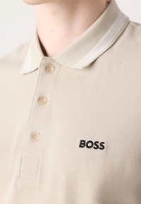 Polo beige à col avec trois boutons et logo noir "BOSS" brodé sur la poitrine gauche, porté par une personne.