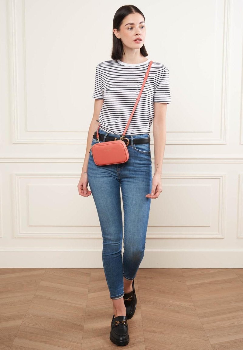 Borsa a tracolla in ecopelle corallo, forma rettangolare con zip dorata. Il modello indossa una maglietta a righe e jeans skinny blu.