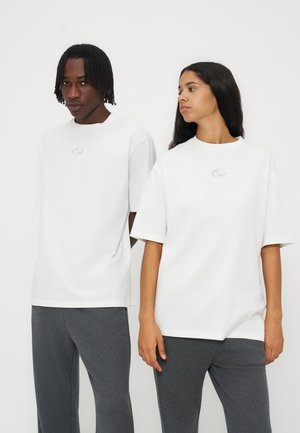 Δύο άτομα φορούν oversized λευκά μπλουζάκια με μικρά λογότυπα, σε συνδυασμό με σκούρα γκρι παντελόνια, στέκονται μπροστά σε απλό φόντο.