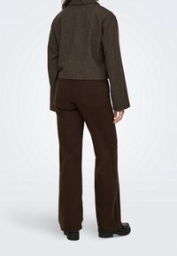 Personne debout, de dos, portant un pantalon droit marron, une veste courte marron, des chaussures noires et une bague à la main droite.