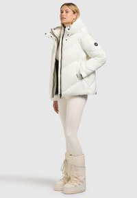 Biała kurtka puchowa z wysokim kołnierzem, zapięciem na suwak i pikowaną strukturą. Noszona na czarnej bluzce i zestawiona z białymi śniegowcami.