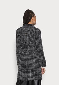 Cappotto in tweed bianco e nero con vita con cintura, silhouette sagomata, dettagli con bottoni e pattina sul retro. Maniche lunghe e colletto.