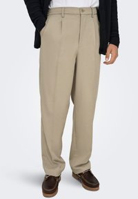 Pantalon plissé beige en tissu lisse, doté de poches latérales et d'une fermeture par bouton, porté avec un cardigan foncé et des chaussures marron.