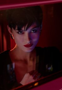 Portrait of a person with short black hair, bold red lips, and silver hoop earrings, against a blurred red background with soft lighting.
