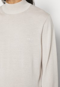 Maglione a collo alto di un beige chiaro realizzato in un morbido tessuto a maglia. Presenta un piccolo logo bianco sul petto e un collo a coste per un tocco di texture.