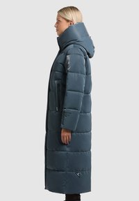 Pitkä, toppatakki tummanturkoosissa värissä, jossa on korkea kaulus ja huppu. Takissa on vetoketjulliset sivutaskut ja helman lähellä oleva logokoriste.