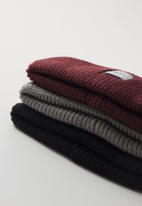 Stack of four knit beanies: burgundy, gray, dark gray, and black. Each has a ribbed texture and a fabric tag on one side.