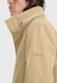 Giacca impermeabile beige chiaro con colletto alto, tasca con zip e logo ricamato sulla manica. Texture liscia, design minimalista.
