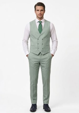 Hombre con traje de tres piezas verde claro, chaleco cruzado, camisa blanca, corbata verde con lunares y zapatos de vestir negros, de pie con las manos en los bolsillos.
