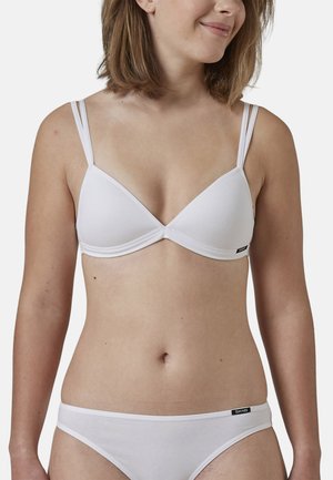 Femme portant un soutien-gorge blanc à double bretelle et une culotte blanche assortie, souriant légèrement, avec des cheveux courts bruns et un fond neutre.